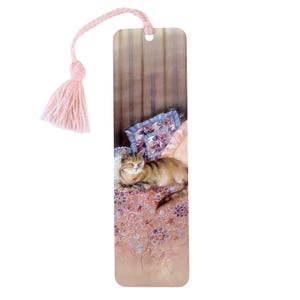 May include: A rectangular bookmark featuring a brown tabby cat resting on a floral patterned surface with a blue pillow. The bookmark has a light pink braided cord with a matching tassel attached through a hole at the top center.