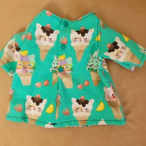 May include: A turquoise fabric garment featuring a repeating pattern of ice cream cones with cat faces, cherries, and strawberries. The garment has a button closure and sleeves. The overall design is whimsical and playful.