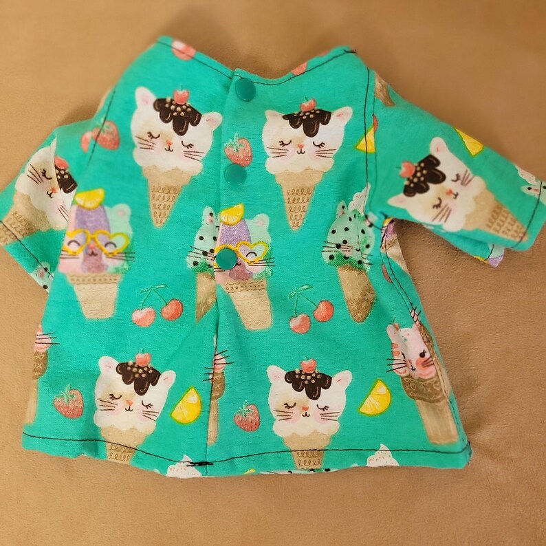 May include: A turquoise fabric garment featuring a repeating pattern of ice cream cones with cat faces, cherries, and strawberries. The garment has a button closure and sleeves. The overall design is whimsical and playful.