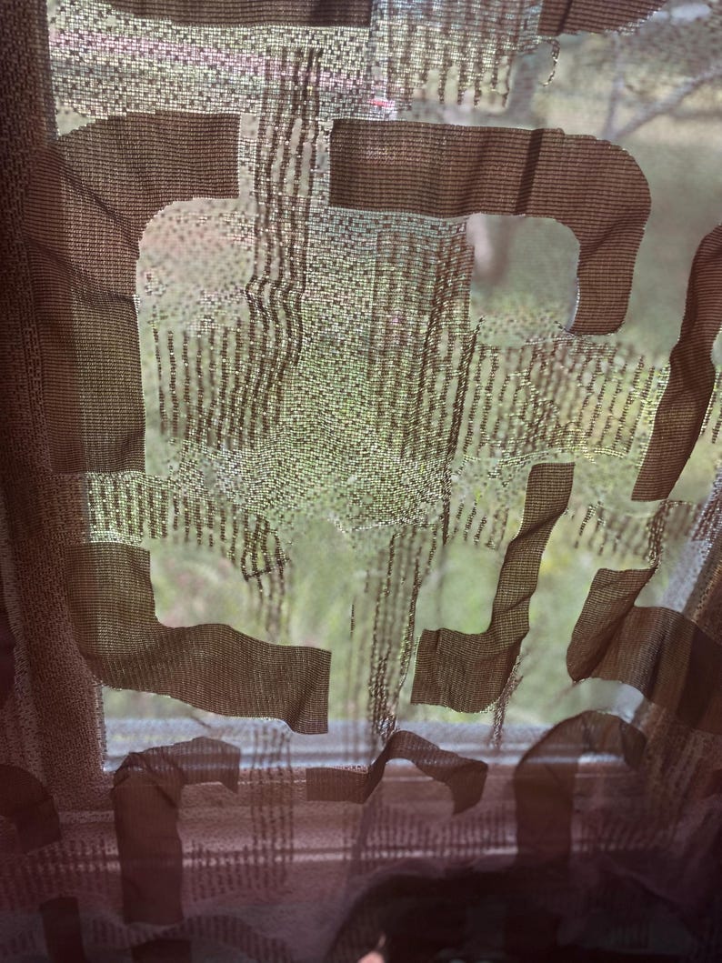 May include: A sheer brown curtain with a large, abstract design. The curtain is made of a lightweight fabric, allowing light to filter through. The design appears to be a series of overlapping shapes, creating a modern aesthetic. The background shows a blurred outdoor scene.
