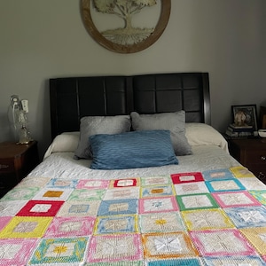 May include: A bed with a black headboard and a white crocheted quilt with colorful squares. The quilt has a variety of colors, including red, yellow, blue, green, and pink. There is a round wooden wall decor with a tree design above the bed.
