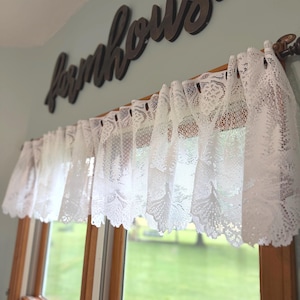 May include: White lace window valance with a floral pattern, hanging above a window. The word "farmhouse" is displayed above the valance in dark brown wooden letters. The window looks out onto a green lawn.