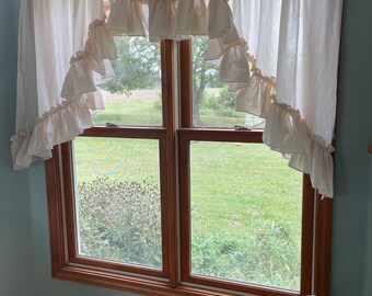 May include: A white ruffled valance with a scalloped edge hangs over a window with a view of a grassy field. The window has a wooden frame and is divided into two panes.
