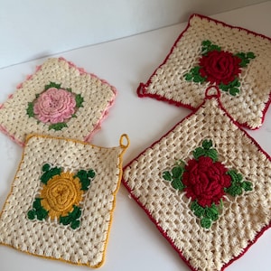 May include: Four square crochet potholders with cream-colored bases. Three feature red roses with green leaves, one features a pink rose with green leaves, and one features a yellow rose with green leaves. Each has a matching colored border and loop.