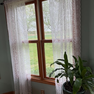 May include: Two white lace curtains with a floral pattern and scalloped edges hang on a wooden rod over a window with a wooden frame. The curtains are sheer and allow light to pass through, revealing a view of green trees and grass outside.