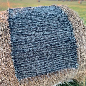 May include: A rectangular, textured rug with a fringe edge, in shades of blue and black, is displayed on a round bale of hay. The rug's design features horizontal stripes. The hay bale is a light brown color, and the background shows a field.