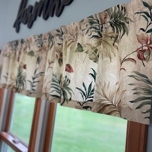 May include: A valance with a tropical leaf pattern in shades of green, brown, and red. The valance is draped over a window, with a wooden frame visible below. The background features a light blue wall and a wooden sign that says "farmhouse".