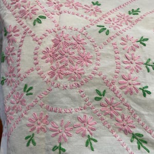 May include: Close-up of white fabric with intricate embroidery. The design features pink floral patterns and green leaf accents. The embroidery is detailed and evenly spaced, creating a textured surface. The overall aesthetic is delicate and handcrafted.
