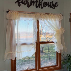 May include: A window with sheer, ruffled curtains and a wooden frame. The curtains are a light cream color, and the word "farmhouse" is displayed above the window in a dark brown script. The window looks out onto a yard.