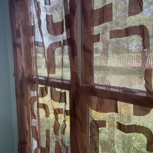 May include: Brown sheer window curtain with a geometric pattern. The curtain is made of a lightweight fabric and hangs in front of a window, allowing light to filter through. The design features abstract shapes.