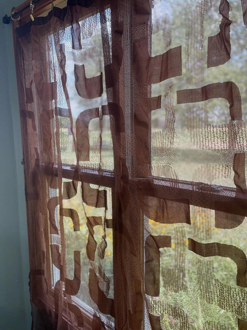 May include: Brown sheer window curtain with a geometric pattern. The curtain is made of a lightweight fabric and hangs in front of a window, allowing light to filter through. The design features abstract shapes.
