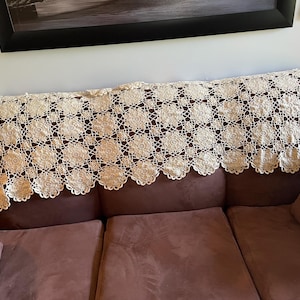 May include: A cream-coloured, crocheted sofa runner with a floral pattern. The runner is draped over the back of a brown sofa. The intricate lace design features repeating circular motifs and a scalloped edge.