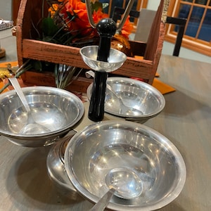May include: A silver three-tiered serving set with a black handle and a loop for carrying. Each tier has a small bowl and a spoon. The set is on a wooden table.