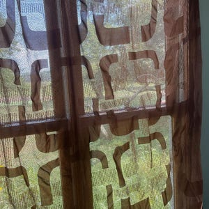 May include: Brown sheer curtains with a repeating geometric pattern. The curtains are hanging in front of a window, allowing natural light to filter through. The pattern is a series of connected, curved shapes.