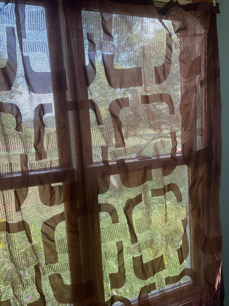 May include: Brown sheer curtains with a repeating geometric pattern. The curtains are hanging in front of a window, allowing natural light to filter through. The pattern is a series of connected, curved shapes.