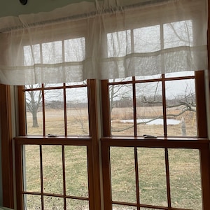 May include: White sheer curtains with ruffles hang in a window with a view of a grassy field and trees in the distance.
