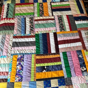 May include: A patchwork quilt featuring a variety of fabric squares and rectangular strips in numerous colors and patterns, including floral, plaid, polka dot, and solid prints arranged in a complex, multi-directional geometric block design.
