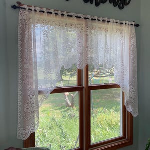 May include: White lace window curtains with floral patterns hang over a window with a view of a green field. A dark brown wooden "farmhouse" sign is mounted above the curtains. The window frame is brown.