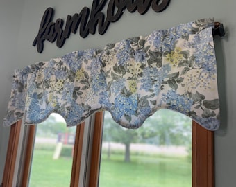 May include: A window valance with a floral pattern in blue, white, and yellow, hangs above a window. The valance is scalloped along the bottom edge. Above the valance, the word "farmhouse" is spelled out in dark wooden letters.