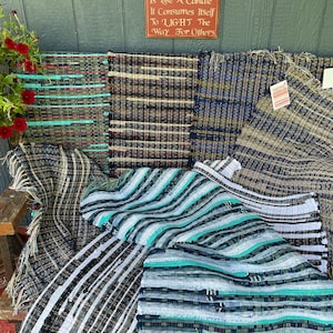 May include: A collection of woven rag rugs in various colours and patterns, including blue, green, brown, and white. The rugs are arranged on a wooden bench with a sign that reads "It is like a candle, it consumes itself to light the way for others."