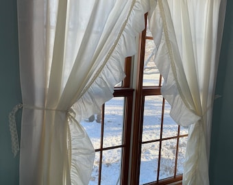 May include: White ruffled curtains with a window view of a snowy yard. The curtains are tied back with white ribbon.