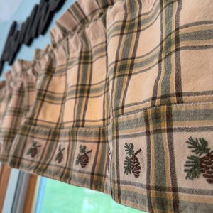 May include: Cream-colored plaid valance with green and brown stripes. The bottom edge features embroidered pinecone designs. The valance is hanging in front of a window with a green view outside.