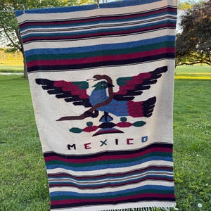 May include: A cream-colored Mexican blanket with a central eagle design in blue, red, and black. The word "MEXICO" is displayed in black letters. The blanket has striped borders in blue, green, and burgundy.