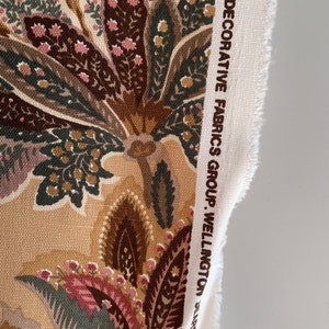 Puede incluir: Primer plano de una muestra de tela con un patr&oacute;n de cachemira en tonos marrones, rosas y verdes. La tela tiene una superficie texturizada y est&aacute; etiquetada como "Decorative Fabrics Group Wellington, Delros Company".