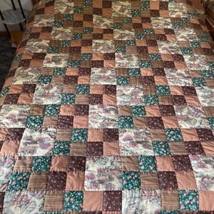 May include: A patchwork quilt featuring a repeating grid of square fabric blocks. The design alternates between solid tan squares, dark brown squares, green floral patterned squares, and light-colored squares depicting small houses and trees.