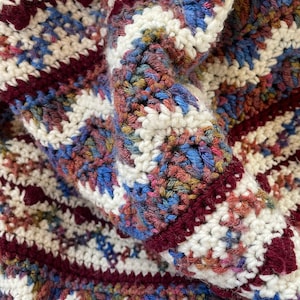 May include: Close-up of a crocheted blanket with a striped pattern in shades of white, burgundy, and blue.