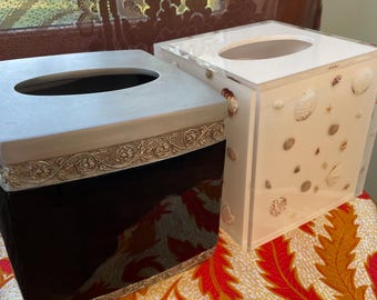 May include: Two decorative tissue box covers. One is black with a silver ornate trim. The other is clear with seashells embedded in the sides. Both have oval openings on top for tissue dispensing.