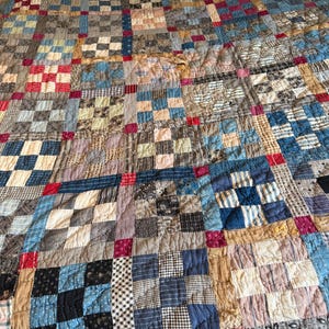 May include: A patchwork quilt featuring a grid of square blocks in various patterns and colors, including blue, tan, brown, and red. The quilt is constructed from multiple fabric squares joined together with small red squares at the intersections.