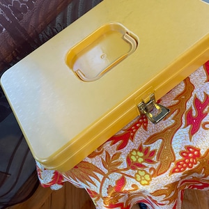 May include: A yellow plastic box with a clear handle and a gold-colored clasp. The box is resting on a table covered with a patterned fabric featuring red, orange, and yellow floral designs.