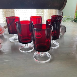 May include: Set of six red glass stemmed drinking glasses with clear bases. The glasses are arranged in a semi-circle on a gray surface.
