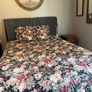 May include: A bed featuring a dark green quilt with a dense floral pattern of pink, red, and cream roses. Two matching square pillows with ruffled edges sit against a dark gray upholstered headboard. A wooden nightstand is positioned to the right.