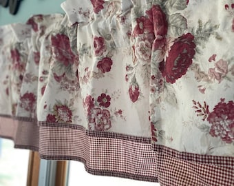 May include: A ruffled window valance with a floral pattern in shades of burgundy, pink, and gray on a cream background. The bottom edge features a burgundy and white gingham trim. The valance is hanging above a window.