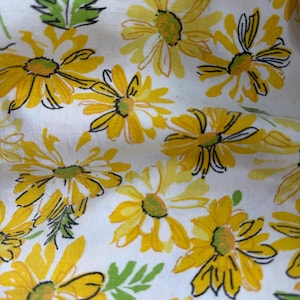 May include: A white fabric featuring a repeating pattern of yellow daisies with green centers and green leaves. The floral design includes black ink outlines that define the petals and foliage, creating a hand-drawn appearance across the textile surface.