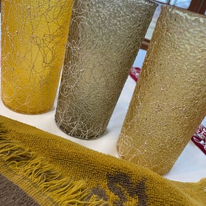 May include: Three textured glass tumblers in yellow, gray, and beige. The glasses have a unique, woven design and are sitting on a brown and yellow fringed towel.