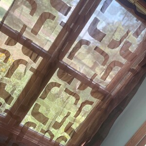 May include: Brown sheer window curtain with a repeating abstract design. The curtain is hanging in front of a window, allowing natural light to filter through. The design is a series of curved shapes.