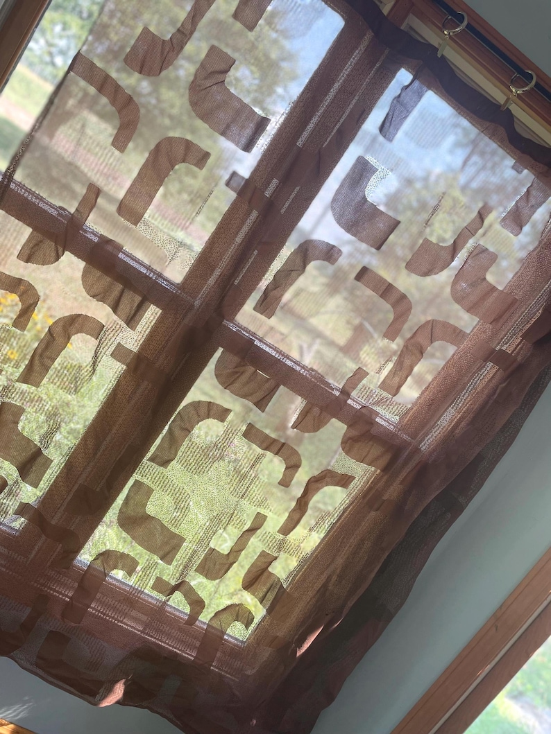 May include: Brown sheer window curtain with a repeating abstract design. The curtain is hanging in front of a window, allowing natural light to filter through. The design is a series of curved shapes.