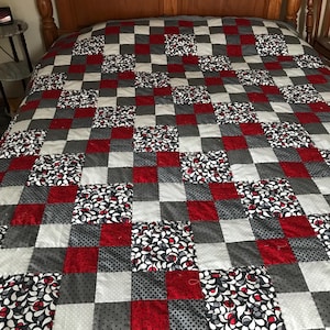 May include: A patchwork quilt in a square pattern, featuring red, grey, white and patterned squares. The patterns include floral and polka dot designs. The quilt is displayed on a wooden bed frame.