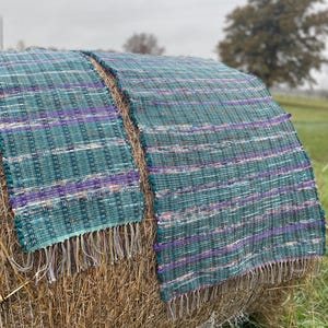May include: Two woven rugs in shades of teal, purple, and white are rolled up on a hay bale. The rugs have a fringed edge.
