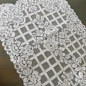 May include: White lace table runner with a floral and geometric design. The runner has a scalloped edge and a delicate, openwork pattern. The lace is intricately detailed, adding a touch of elegance to any table setting.