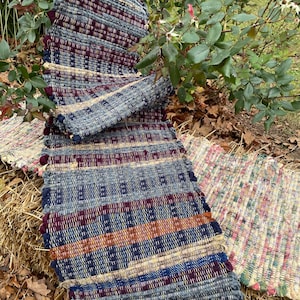 May include: Two woven rugs with a fringe. The rug on top is blue, red, orange, and yellow. The rug on the bottom is multi-colored with a green, pink, and white pattern.