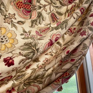 May include: A cream-colored fabric with a floral pattern in shades of red, pink, green, and yellow. The fabric has a gathered or ruffled look, and the floral pattern is intricate and detailed.