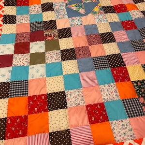 May include: A colourful patchwork quilt with a variety of fabrics and patterns. The quilt is made up of squares in different colours, including floral, striped, and solid fabrics. A central pinwheel design is a focal point, and a red and white chevron border.