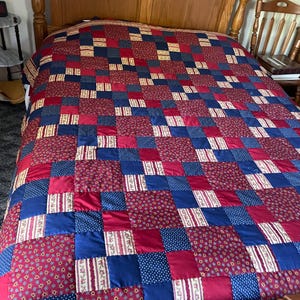 May include: A patchwork quilt in shades of red, navy blue, and cream covers a wooden bed frame. The quilt features a repeating pattern of squares and rectangles, creating a classic, cozy aesthetic. The quilt's design includes floral and polka dot patterns.