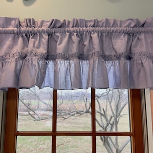 May include: A light blue ruffled window valance. The valance is made of a soft fabric and has a gathered top and a ruffled bottom edge. The valance is hanging above a window with a view of a tree and a field.