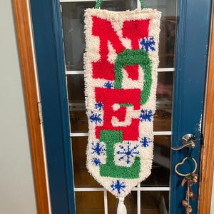 May include: A handmade Christmas wall hanging with the word "NOEL" in red and green, accented with blue snowflakes on a cream background. It has a white tassel and a green hanging cord.