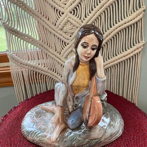 May include: A ceramic figurine of a woman sitting with a guitar. She is wearing a blue and yellow outfit and has brown hair. The figurine is sitting on a base that looks like a grassy area.